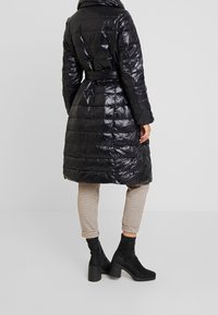 Abrigo acolchado negro y brillante con cintura ceñida, cuello alto y diseño acolchado. Llevado sobre pantalones de cuadros y botines negros.