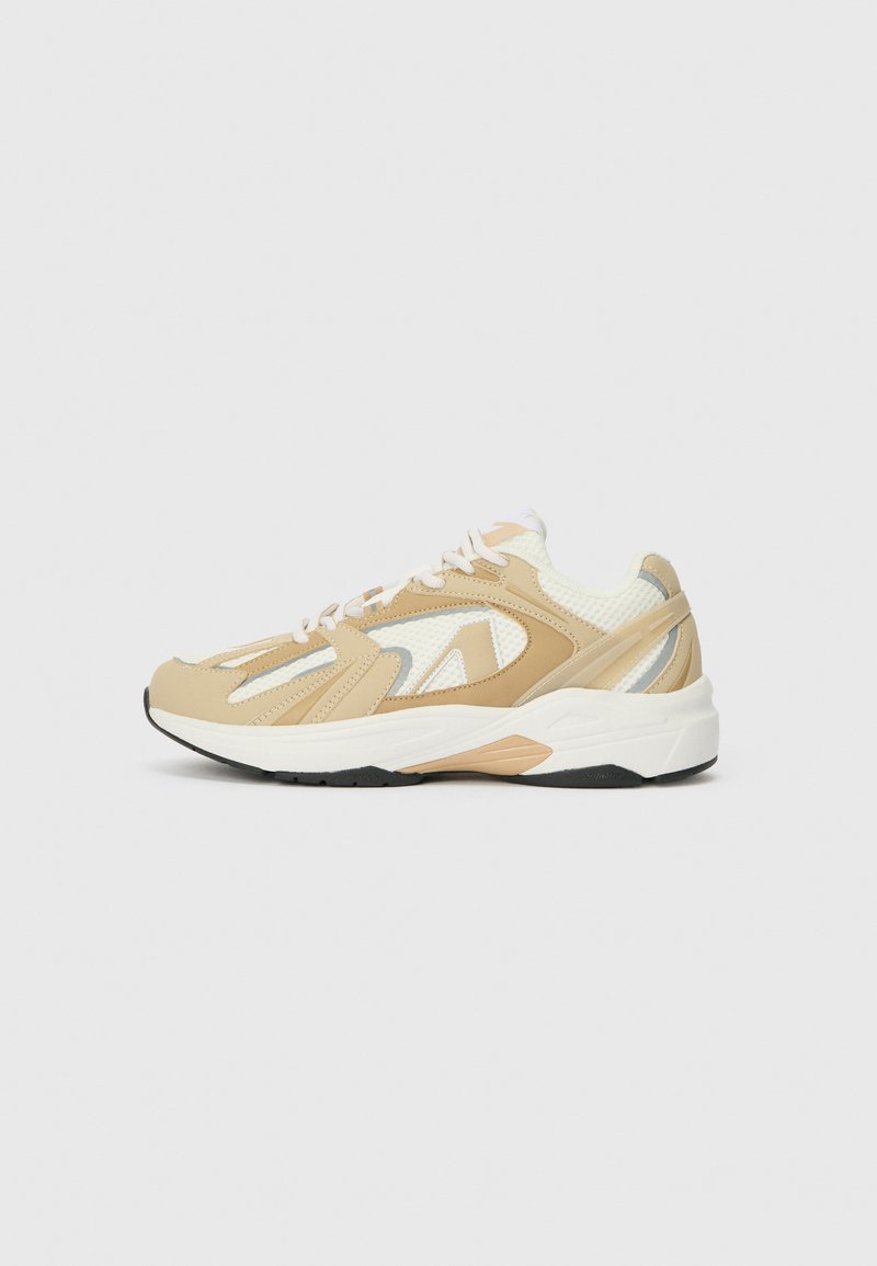 Zapatilla deportiva beige con parte superior de malla y sintético, con detalles en gris claro, cordones y una suela texturizada. Diseño redondeado, talón acolchado.