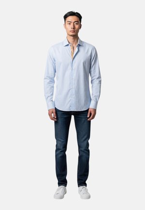 Uomo in piedi che indossa una camicia azzurro chiaro con bottoni, jeans blu scuro e scarpe da ginnastica bianche su uno sfondo chiaro e semplice.