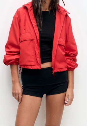 Femme portant une veste rouge courte à capuche et un top court noir avec un short noir assorti, debout devant un fond blanc uni.