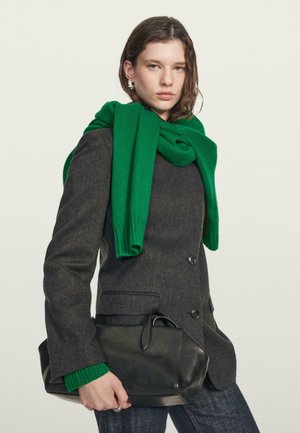 Blazer grigio in lana con un design aderente e sciarpa in maglia verde drappeggiata sopra. Il modello tiene una borsa in pelle nera con bordi arrotondati.