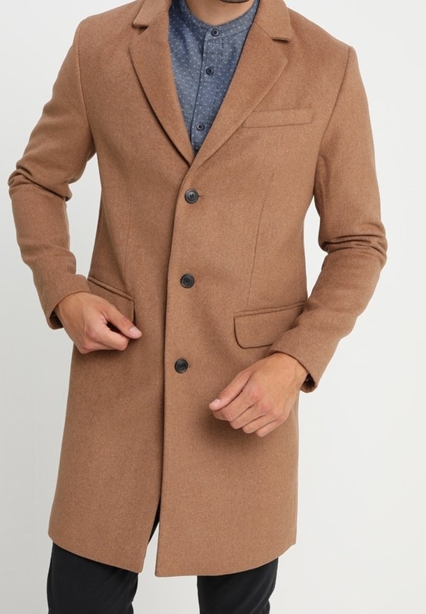 Cappotto in misto lana marrone con colletto a punte, due tasche frontali e tre bottoni neri. Indossato sopra una camicia blu con motivo.