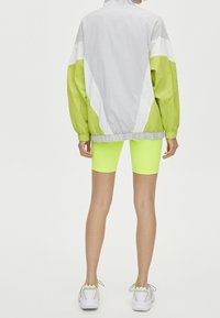 Giacca a vento leggera grigia con dettagli verdi, caratterizzata da un colletto alto e una vestibilità oversize. Abbinata a pantaloni da bicicletta giallo fluorescente e sneaker bianche.
