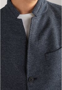 Navy textured jacket with a stand-up collar, front button closure, and a chest pocket. Fabric appears thick and structured.