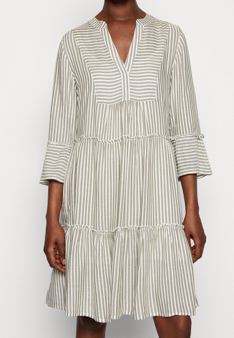 Femme portant une robe ample longueur genoux avec des rayures verticales beiges et blanches et des manches cloche 3/4.