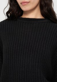 Femme aux longs cheveux foncés portant un pull en maille noire texturée, révélant le col et le haut de la poitrine.