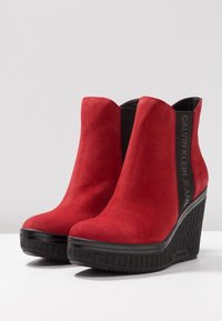 Czerwone zamszowe botki na koturnie, z czarną teksturowaną platformą i elastycznymi panelami po bokach. Logo widoczne z boku.