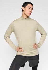 Μπεζ αθλητικό μακρυμάνικο τοπ με φερμουάρ μέχρι τον λαιμό, με διακριτική τονική σήμανση στους ώμους, σε συνδυασμό με μαύρο σορτς. Λεία, εφαρμοστή υφή.