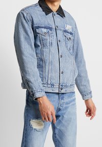 Ljusblå denimjacka med svart krage, två bröstfickor, tryckknappar och sliten textur, kombinerad med matchande trasiga jeans.