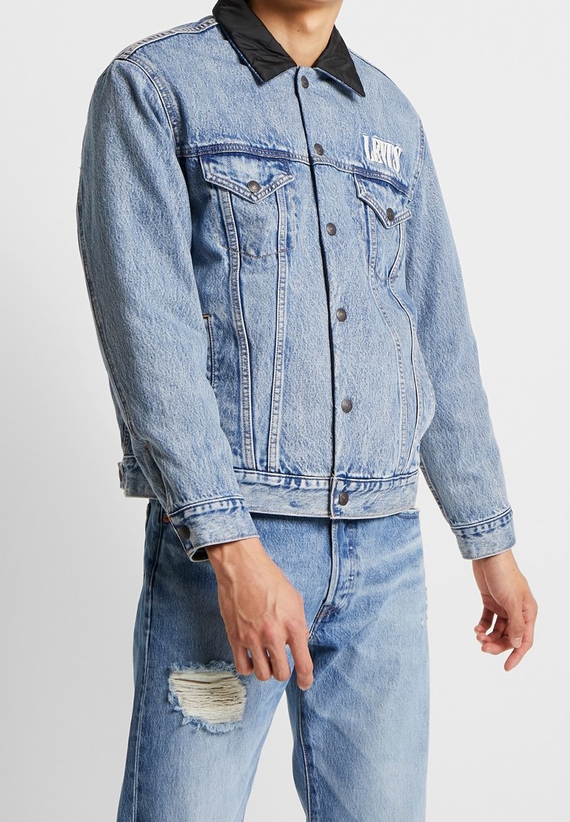 Ljusblå denimjacka med svart krage, två bröstfickor, tryckknappar och sliten textur, kombinerad med matchande trasiga jeans.