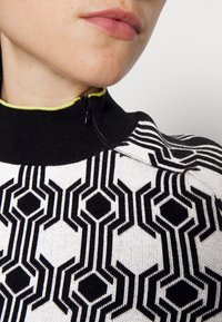 Jersey de punto con un cuello alto negro, base blanca y un audaz patrón geométrico hexagonal en blanco y negro, acentuado con una cremallera verde.