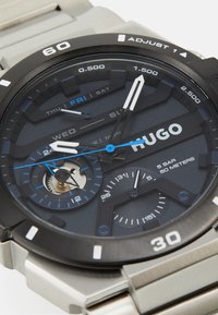 Hugo-Uhr mit einem dunkelblauen strukturierten Ziffernblatt, Gehäuse und Armband aus silbernem Edelstahl. Zu den Funktionen gehören ein Datum- und Wochentagsanzeige, zwei kleine Zifferblätter und markante Indizes.