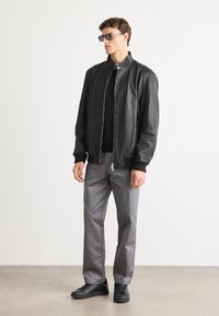 Hombre con chaqueta de cuero negra, camiseta negra, pantalones grises, zapatillas negras y gafas de sol, de pie contra un fondo claro y liso.