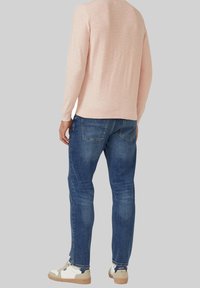 Persoon draagt een lichtroze top met lange mouwen, blauwe denim jeans en witte sneakers, staat met de rug naar de kijker toe.