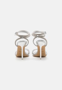 Sølv høyhælte sandaler med et buet stroppdesign, metalliske detaljer og en teksturert stropp rundt ankelen. Klare stilettihæler.