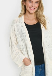 Lätt creamfärgad cardigan i öppen stickningsmönster, lång ärm och fickor; bärs över en svart topp med en avslappnad passform.