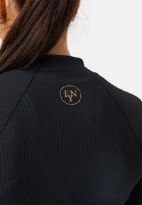 Svart athletic top med ribbad textur, hög krage och en cirkulär gold-logotyp på baksidan med bokstäverna "ENY".