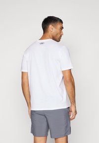 T-shirt blanc à manches courtes avec un col ras du cou, arborant un petit logo noir au dos. Shorts gris visibles, le tissu semble léger et respirant.