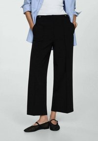 Pantalons larges noirs en tissu lisse, avec des plis à l'avant et des accents en métal doré. Associés à un haut blanc et une chemise bleue.