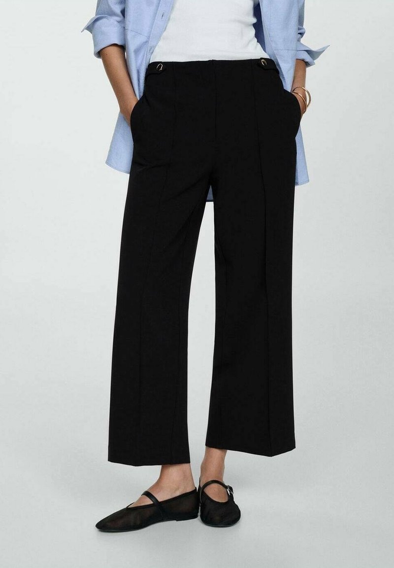 Pantalons larges noirs en tissu lisse, avec des plis à l'avant et des accents en métal doré. Associés à un haut blanc et une chemise bleue.