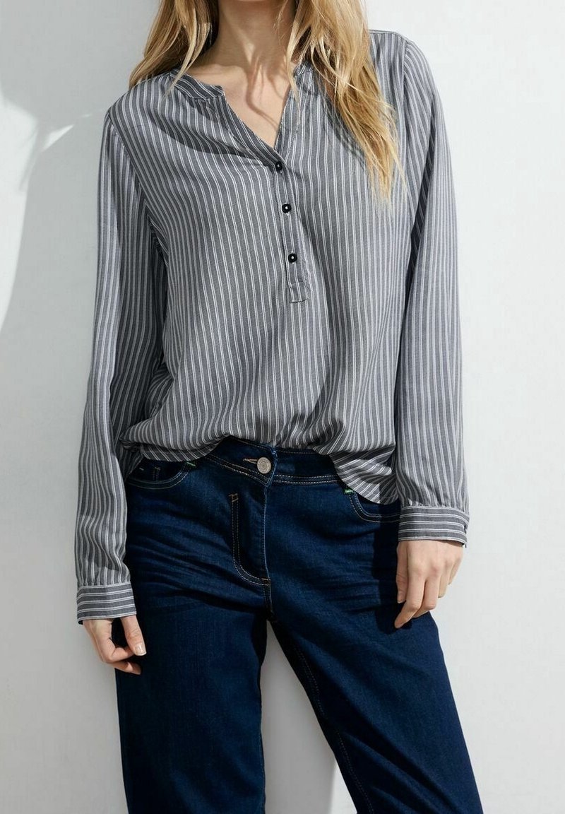 Gestreepte grijze blouse met lange mouwen, voorzien van een V-hals en zwarte knopen, gecombineerd met donkerblauwe jeans.