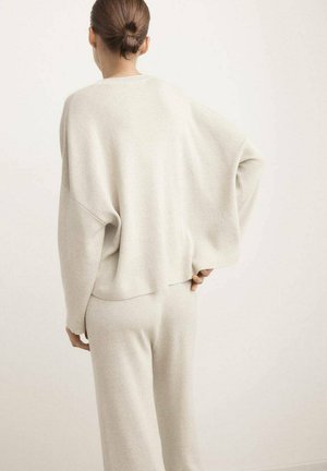 Mujer con el cabello recogido en un moño, vistiendo un suéter de punto holgado color beige claro y pantalones anchos a juego, de pie con una mano en la cintura.