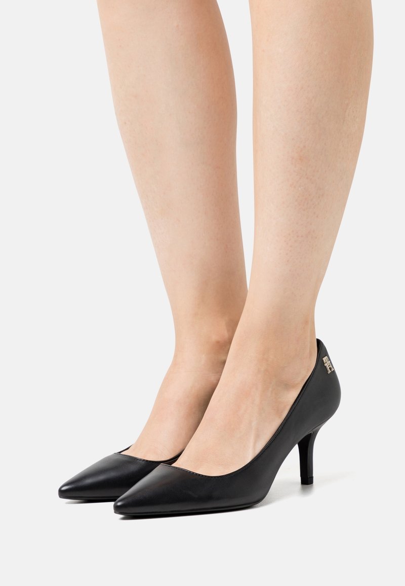 Jambes portant des chaussures à talons hauts noires à bout pointu avec un petit logo en métal sur le côté extérieur, debout sur un fond blanc.
