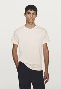 Giovane uomo con capelli corti e scuri che indossa una t-shirt bianca semplice e pantaloni neri, in piedi contro uno sfondo chiaro.