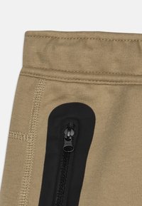 Béžové sportovní kalhoty s elastickým pasem, boční kapsou na zip v černé barvě a viditelnými detaily švů. Měkká, pružná látka.