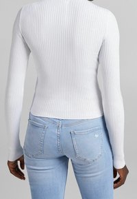 Person i en vit ribbad långärmad tröja och ljusblå denimjeans, sedd bakifrån mot en enfärgad bakgrund.