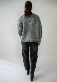 Grijze gebreide trui met een gestructureerd patroon, gecombineerd met losse zwarte jeans en zwarte enkellaarsjes met hak. Uitzicht van achteren.