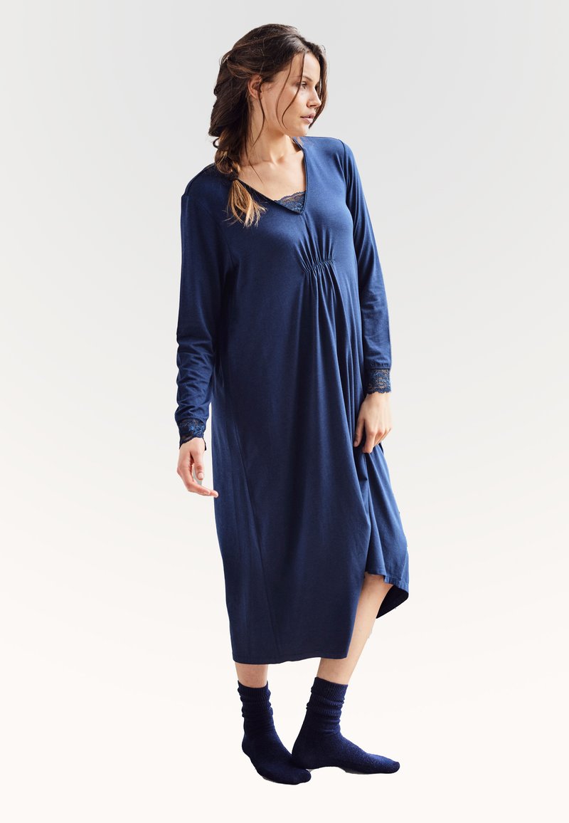 Marineblå langærmet natkjole med blondekant ved halsudskæringen og ærmerne, med en samlet bøst og knælang kant. Båret med mørke sokker.