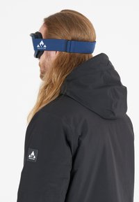 Hiihtolasit, joissa on sininen hihna, johon on merkitty vuorilogot, musta kehys ja vaahtomuovipehmustus, joita käyttää henkilö mustassa hupparissa.