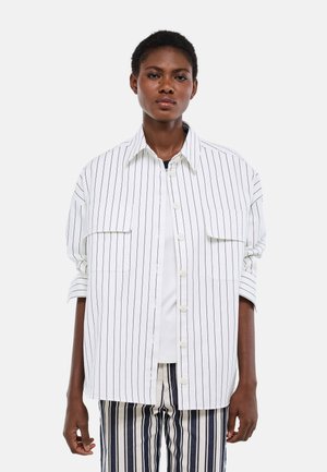 Femme portant une chemise blanche boutonnée à fines rayures noires verticales et un pantalon assorti à rayures verticales, debout devant un fond uni.