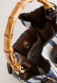 Sac à main avec poignée en bambou, extérieur tissé bleu marine et blanc, garniture en cuir marron et intérieur ouvert montrant une poche zippée et un fermoir magnétique.