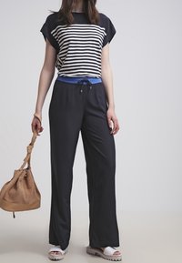 Woman wearing black wide-leg pants with blue waistband, black and white striped top, white sandals, holding tan handbag with perforated design.