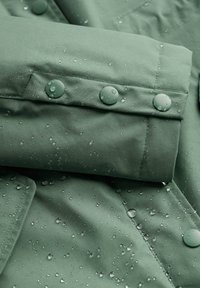 Voděodolná zelená tkanina s hladkým povrchem, opatřená dvěma matnými zelenými patentkami a kapkami vody na povrchu.