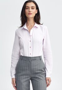 Jasnoróżowa koszula zapinana na guziki z długimi rękawami, z prostym kołnierzykiem i małymi guzikami. W zestawie z szarymi spodniami w paski.