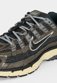 Αθλητικό παπούτσι Nike με μαύρο δίχτυ, γκρι αντανακλαστικές επικαλύψεις, ανοιχτό γκρι κορδόνια και κρεμ υφή στη σόλα, σε κλίση για να δείχνει πλευρικές και πάνω λεπτομέρειες.