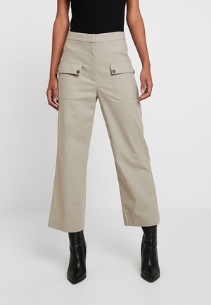 Woman wearing beige wide-leg pants with large front flap pockets and black heeled ankle boots against a plain background.