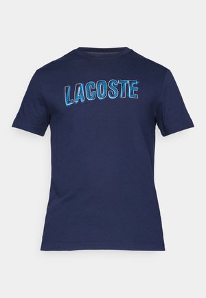 T-shirt in cotone blu navy con scollatura a girocollo e maniche corte, caratterizzata dalla scritta "LACOSTE" stampata in grandi lettere blu in grassetto sul davanti.