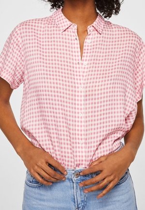 Nainen, joka pukeutunut lyhythihaiseen vaaleanpuna-valkoraidalliseen napitettuun paitaan, joka on työnnetty vaaleansinisten farkkujen sisään, kädet vyötärön lähellä.