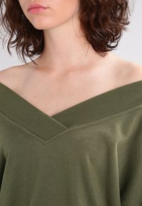 Olivgrön topp med avskuren axel och djup V-ringning samt ribbad kant. Mjukt tyg med en avslappnad passform och inga märkbara mönster.