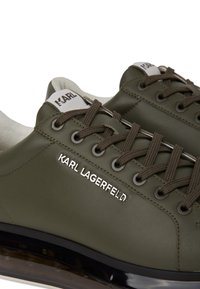 Sneaker in pelle verde oliva con punta rotonda, lacci piatti tono su tono, logo argentato e fodera in vista. Suola traslucida nera con dettaglio di ammortizzazione.