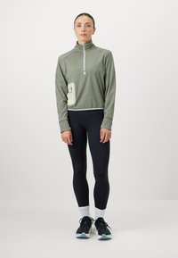 Casaco de meia zip verde com padrão, colarinho alto e um bolso lateral, combinado com leggings pretas e sapatilhas de desporto. Textura suave e design ajustado.