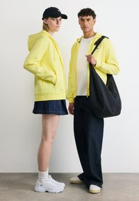 Deux personnes portant des sweat-shirts à capuche jaunes ; la personne à gauche porte une casquette noire, une jupe en jean et des baskets blanches, la personne à droite tient un grand sac noir.