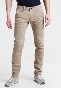 Pantalons en coton beige à coupe slim, avec poches avant et arrière et fermeture à boutons. Portés avec des baskets gris clair.