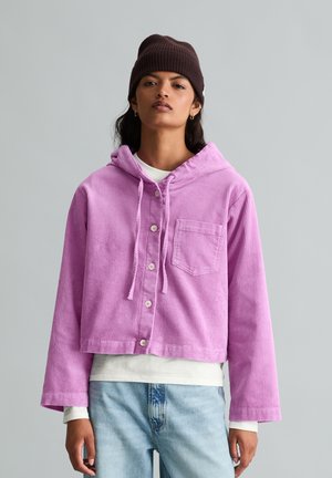 Jeune femme portant un bonnet marron, un sweat à capuche en velours côtelé lavande boutonné, une chemise blanche et un jean bleu clair, debout devant un fond uni.