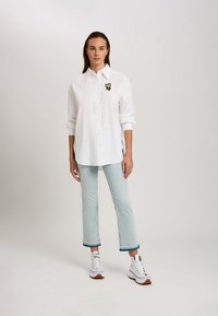 White button-up shirt with long sleeves and a black embroidered logo on the chest. Paired with light blue cropped jeans and white sneakers.