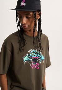 Camiseta verde oliva con un diseño gráfico que presenta una criatura azul y texto en rosa. Gorra negra con logo de rosa rosa. Peinado trenzado.